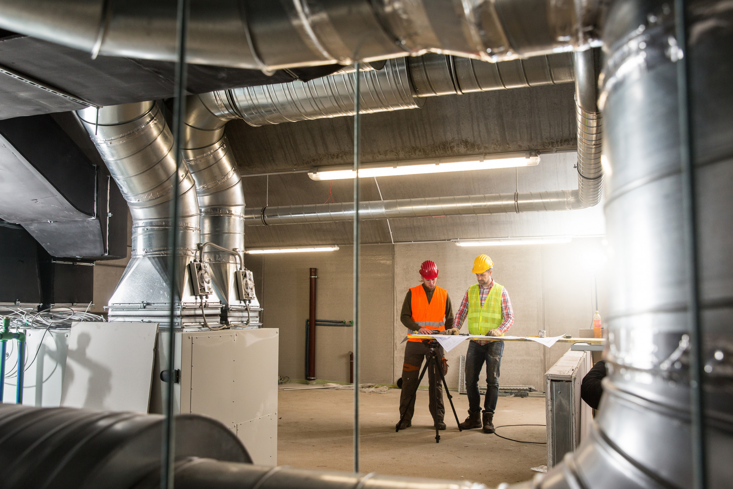 Two construction workers in safety vests and hard hats review blueprints inside a large industrial HVAC system with ductwork overhead.