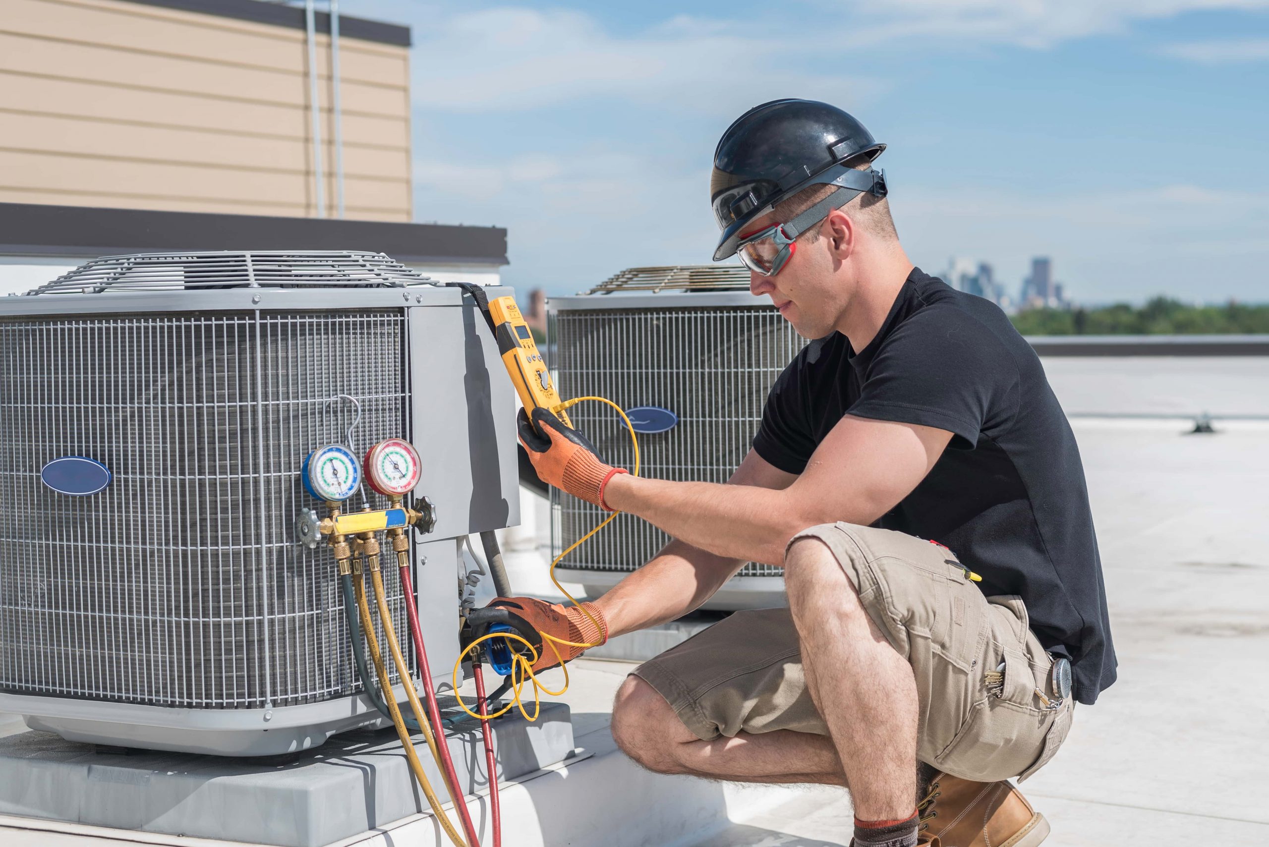 A technician in a black hard hat and safety glasses inspects a rooftop HVAC unit with a digital meter and pressure gauges.