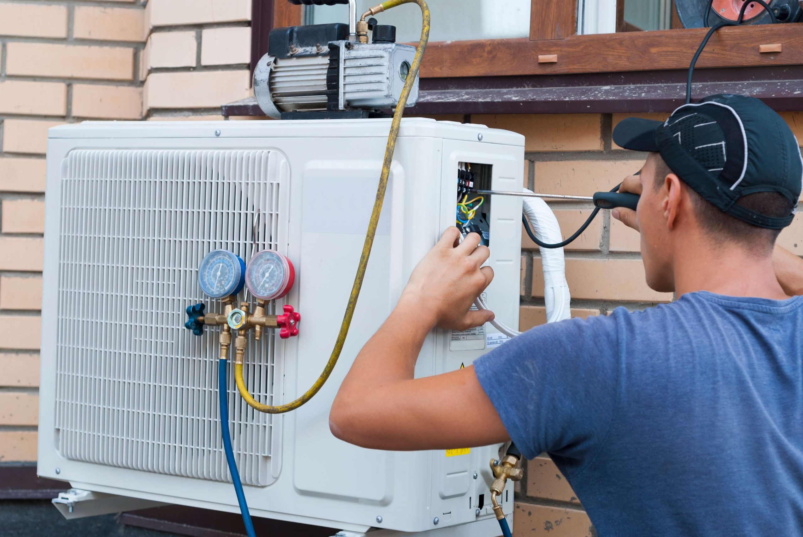 seattle heating cooling services by product lennox Bellevue scaled - Seattle Heating & Cooling A technician in a blue shirt repairs an outdoor air conditioning unit mounted on a brick wall, using a screwdriver and gauge set.