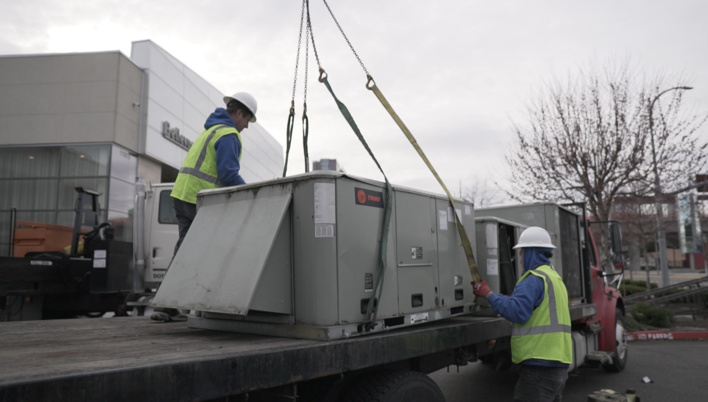 seattle heating cooling services cooling services bellevue - Seattle Heating & Cooling Two HVAC installers secure a large Trane rooftop unit onto a flatbed truck using straps and a crane outside a modern facility.