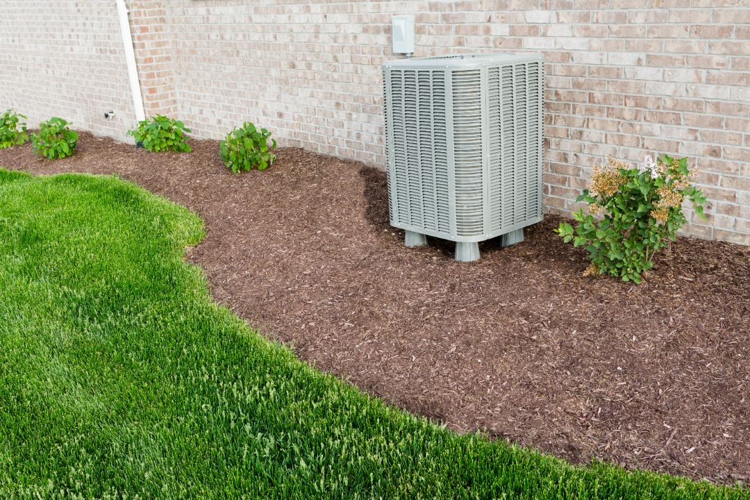 Outdoor air conditioning unit installed against a brick wall, surrounded by mulch, green grass, and neatly trimmed bushes.