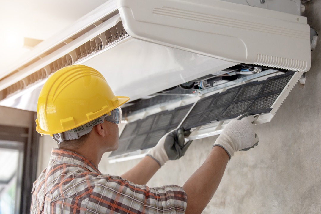 HVAC technician in a yellow hard hat and safety goggles services a wall-mounted air conditioning unit, inspecting and cleaning the filter.