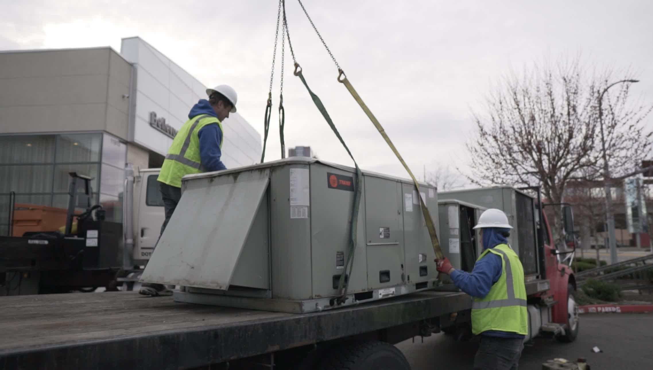 seattle heating cooling redmond refrigeration services furnace installation - Seattle Heating & Cooling Workers in safety vests guide a large rooftop HVAC unit being hoisted onto the flatbed truck outside a commercial building.